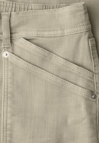 Gros plan sur un pantalon en denim beige montrant la poche avant, les rivets, la passants de ceinture et les coutures détaillées.