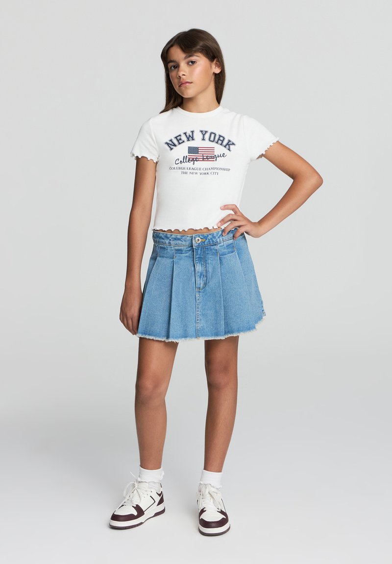 T-shirt bianco corto con stampa "NEW YORK", abbinato a una gonna di jeans blu a vita alta con plissettature e orlo sfilacciato. Sneakers bianche.