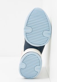 Semelle d'une basket avec une bande de roulement en caoutchouc bleu clair présentant des rainures horizontales et le logo "Michael Kors" sur une semelle intermédiaire marine.