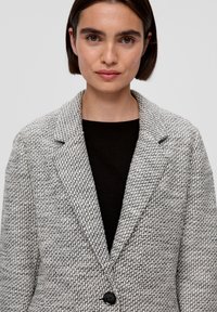 Strukturiertes graues und weißes Blazer mit schwarzem Knopf, Reverskragen und offenem Vorderteil; getragen über einem schwarzen Oberteil mit strukturiertem Design.
