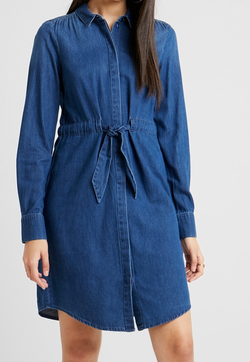 Abito camicia in denim blu scuro, con fronte a bottoni, maniche lunghe, una cintura da annodare in vita e un orlo curvo.