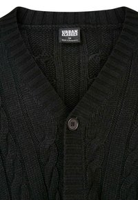Svart kabelstrikket cardigan med V-hals, ribbestrikket krage og en enkelt mørk knappfeste. Lappen viser "Urban Classics" og størrelse M.