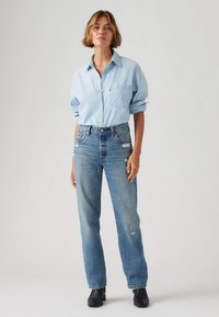 Chemise en denim bleu clair avec des manches retroussées, devant à boutons, et une poche sur la poitrine. Portée avec un jean bleu délavé taille haute et des bottines noires.