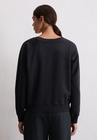 Femme aux cheveux bruns tirés en arrière, portant un sweat-shirt noir à manches longues simple et un pantalon noir, se tenant face à un mur texturé blanc.
