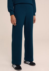 Teal, strukturerade, långa byxor med sidofickor, i kombination med svarta sneakers med vita ränder. Avslappnad, rak design.