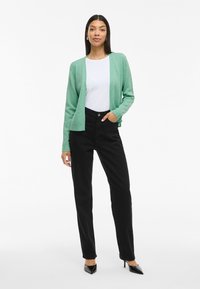 Cardigan de malha verde sobre uma t-shirt branca, combinado com calças pretas de cintura alta. A mulher usa sapatos de salto agudo pretos e brincos em argola.
