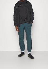 Černá zipová bunda přes černý top, doplněná tyrkysovými tepláky a černými kopačkami. Obsahuje loga Nike a elastick�é manžety na kalhotách.