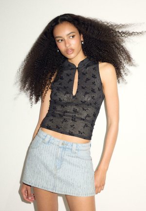 Ung kvinde med fyldigt krøllet hår, iført en sort mønstret ærmeløs top og en lyseblå denim mininederdel mod en ensfarvet baggrund.