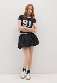 Maglietta nera corta con bordo bianco e numero "91" argentato sbiadito, abbinata a una gonna mini A-line nera e voluminosa. Sneakers bianche.