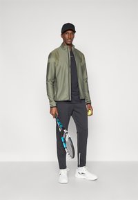 Αθλητική εμφάνιση που περιλαμβάνει ένα ελαιόχρωμο μπουφάν με υφασμάτινες λεπτομέρειες στους ώμους, μαύρη μπλούζα, γκρι παντελόνι και λευκά sneakers, κρατώντας μια ρακέτα και μια μπάλα.