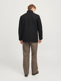 Crni kaput s visokim ovratnikom, teksturiranog materijala, ravnog kroja i proreza straga, uparen s bež hlačama i crnim cipelama.