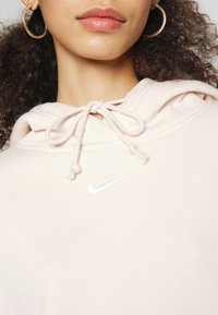 Sudadera de color beige claro con cuello con cordón, presenta un logo blanco de Nike, material de algodón suave y un corte holgado. Detalle en primer plano del cuello.