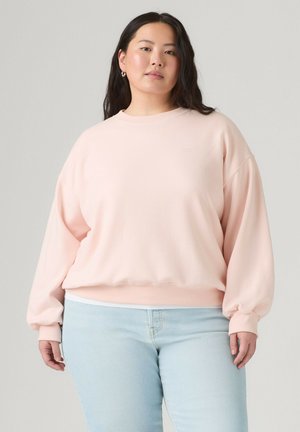 Ung kvinde med langt mørkt hår iført en lyserød oversized sweatshirt og lyseblå jeans, stående mod en grå baggrund.