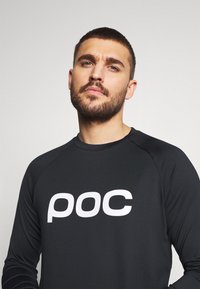 Svart långärmad skjorta i slät tyg, med en djärv vit "poc" logotyp på framsidan och raglanärmar för enkel rörelse.
