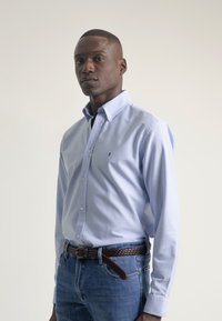 Camisa de botón de color azul claro con cuello y logo bordado, combinada con jeans de mezclilla azules y un cinturón trenzado marrón.