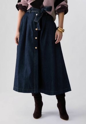 Donna con gonna midi in denim scuro con bottoni frontali e cintura annodata in vita, maglione multicolore, braccialetti d'oro e stivali in suede neri.