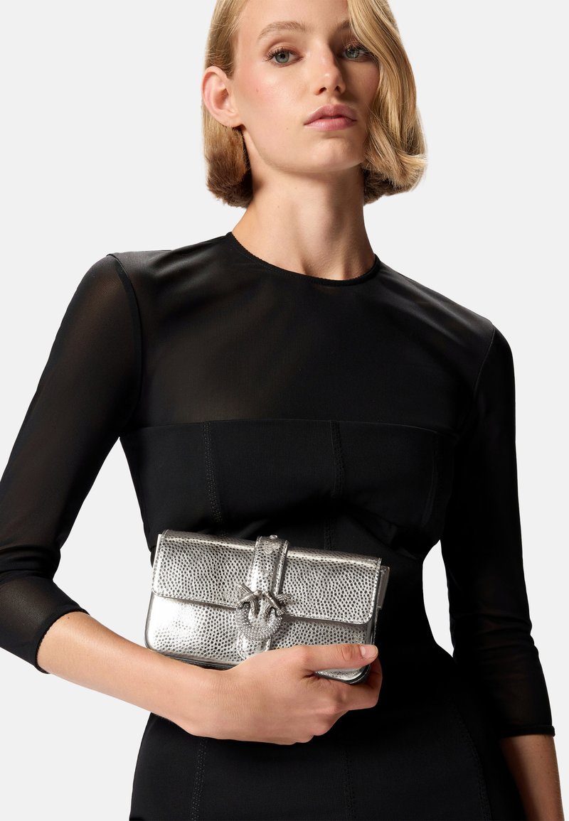 Borsa a pochette metallica argento con superficie texturizzata e chiusura a forma di fiocco, tenuta in mano davanti a un vestito nero aderente con maniche lunghe.