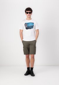 Hvit T-skjorte med blå grafisk design, olivengrønne cargo shorts, svarte sokker og svarte sko; modellen står med armene langs siden.