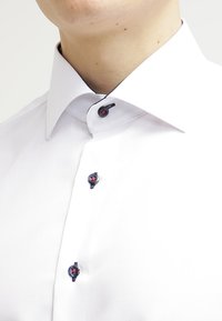 White dress shirt with a button-down collar, featuring dark blue buttons with red accents. Smooth texture and tailored fit.