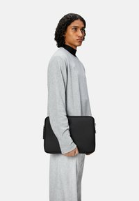 Sort lynlås laptop taske holdt af en person, lavet af glat læderlignende materiale. Simpel form uden mønstre, parret med gråt tøj.