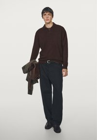 Mörkbrun sweater med krage, tillsammans med mörka byxor och ett bälte. Modellen håller en brun ytterplagg. Slät textur, minimalistisk design.