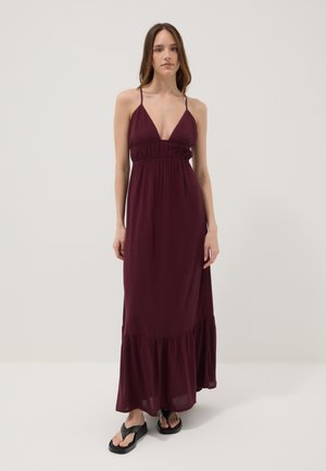 Femme portant une robe maxi bordeaux sans manches avec fines bretelles et des sandales à plateforme noires, debout devant un fond clair uni.