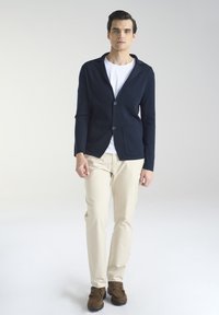 Granatowy kardigan z wyciętym kołnierzem, dwoma przednimi kieszeniami i zapięciem na guziki, noszony na białej koszulce, zestawiony z beżowymi spodniami i brązowymi butami.