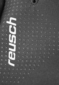 Surface textile noir avec un motif en relief en forme de croix, comportant un logo "reusch" blanc proéminent dans une police moderne.