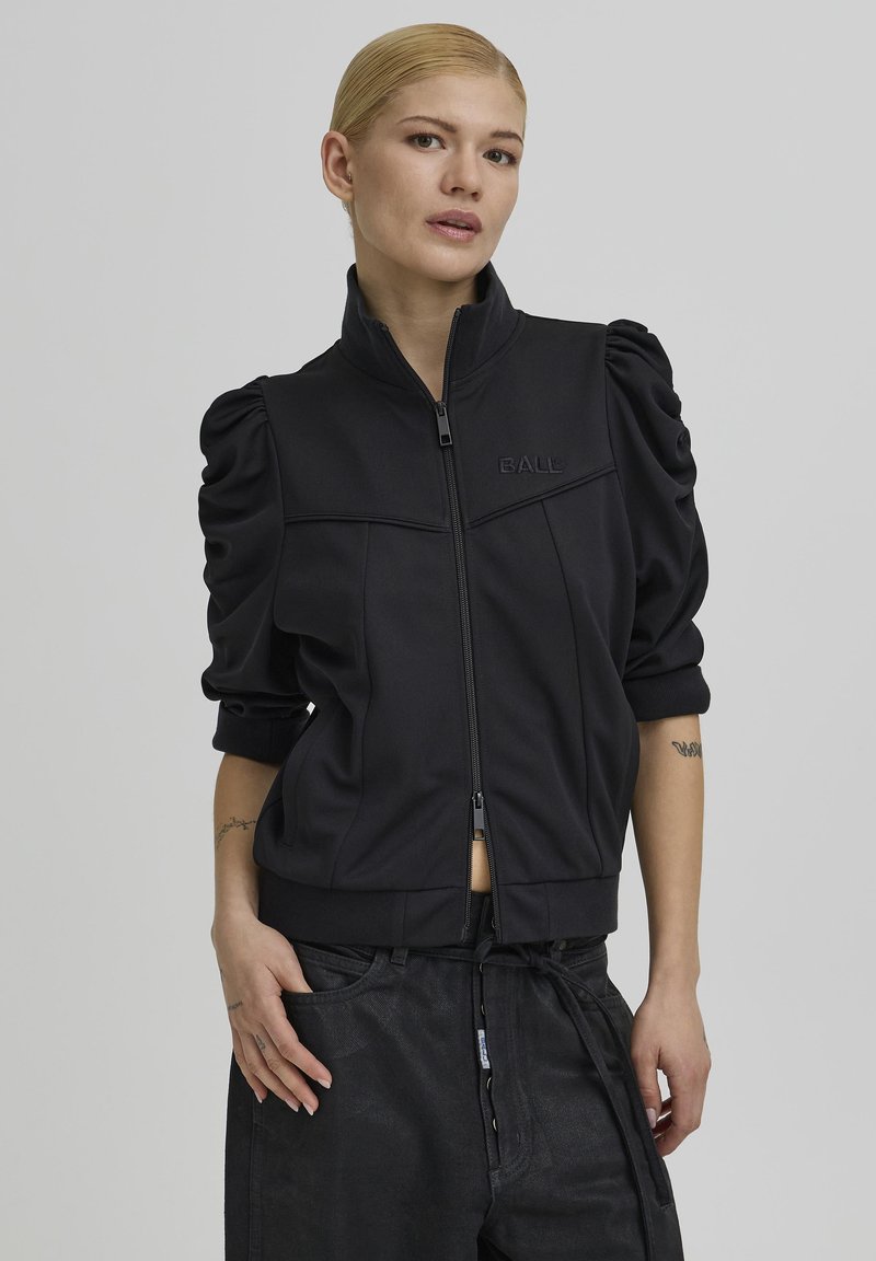 Crna jakna na patentni zatvarač s napuhanih rukava, glatkog tkanja i suptilnim logotipom. Uparena s širokim tamnim hlačama za ležeran izgled.