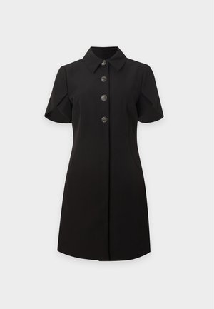 Mujer con vestido negro de manga corta con botones decorativos, de pie, combinado con tacones slingback negros y grandes pendientes de aro.