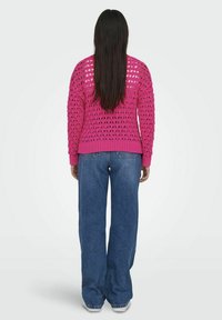 Roze gebreide trui met grote uitsnijdingen, geribbelde manchetten en zoom, gecombineerd met wijde blauwe spijkerbroek en witte schoenen, gezien van achteren.
