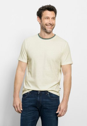 Uomo sorridente, indossa una maglietta crema a maniche corte con bordo verde scuro al colletto e jeans blu, in piedi contro uno sfondo chiaro e uniforme.