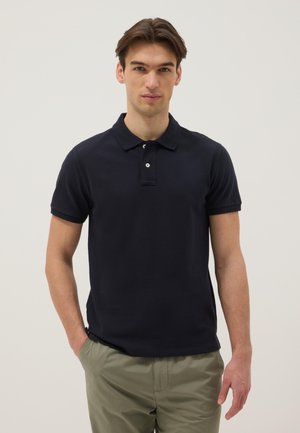 Junger Mann, der ein schwarzes, kurzärmeliges Poloshirt und olivgrüne Hosen trägt, steht mit einer Hand in der Tasche vor einem schlichten Hintergrund.