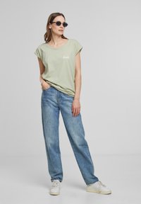 T-shirt vert clair à manches courtes avec le texte "forêt", associé à un jean bleu ample et des baskets blanches. Tissu doux et coupe décontractée.