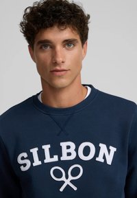 Sudadera azul marino de algodón con cuello redondo. Presenta bordado en blanco con la inscripción "SILBON" y un logo de tijeras en el frente.