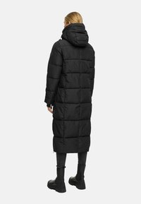 Femme vue de dos portant un long manteau matelassé noir avec capuche, un pantalon noir et des bottes noires chunky sur un fond uni.