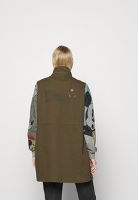 Olijfgroene vest met een hoge kraag, een gestructureerde stof en getailleerde sweatshirtmouwen met kleurrijke cartoonfiguren.