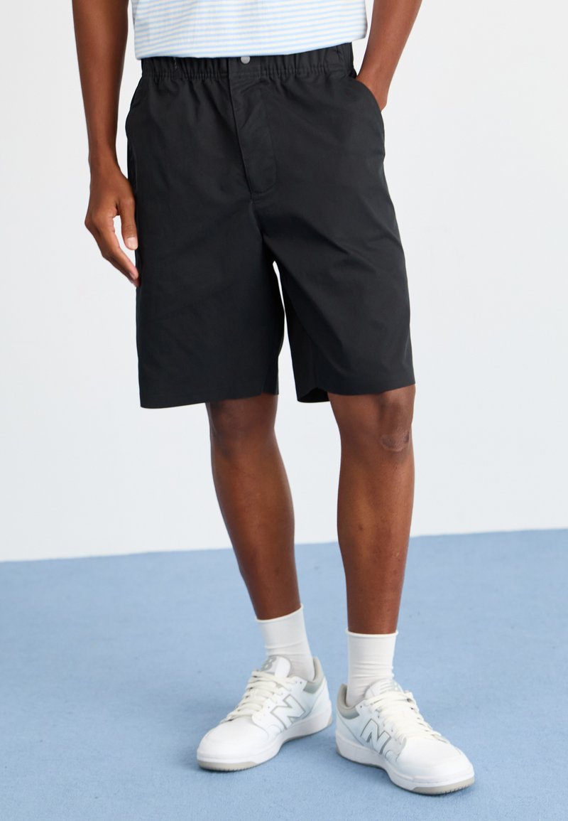 Svarte shorts med elastisk midje og lommer, sammen med hvite joggesko og lyse sokker. Enkel, strømlinjeformet design.