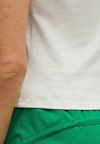 Valkoinen lyhythihainen paita, jossa on teksturoitu pinta ja pieni brodeerattu logo alakulmassa, yhdistettynä vihreisiin urheilushortseihin.