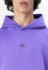 Lila Kapuzenpullover aus weichem Stoff, mit einem metallischen Logo-Detail auf der Vorderseite. Das Design umfasst eine Kängurutasche und eine verstellbare Kordelzugkapuze.