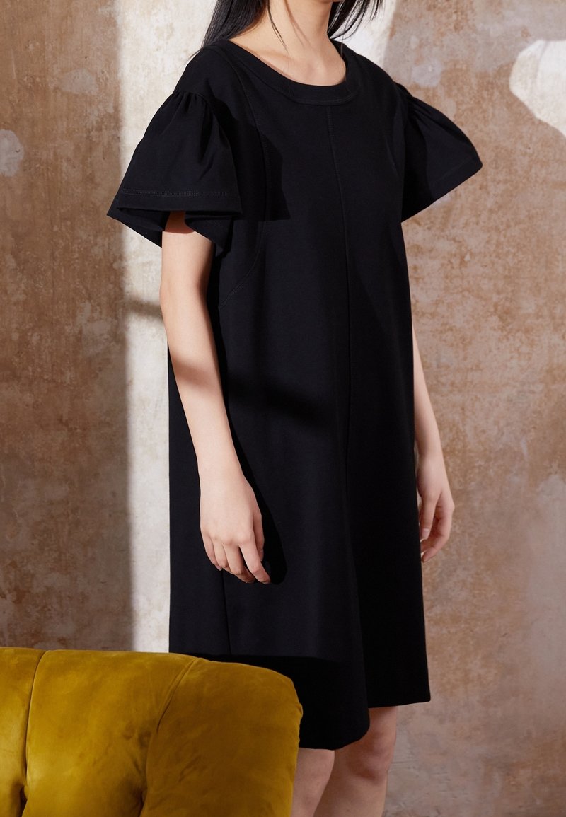 Femme portant une robe noire à larges manches courtes à volants, debout près d'une chaise jaune en velours contre un mur beige texturé.