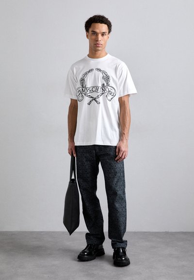 T-shirt blanc Moschino avec un design graphique noir et une coupe oversized, associé à un pantalon noir à motifs et des chaussures noires brillantes, portant un sac fourre-tout.