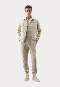 Beige, quiltet vest over en matchende tracksuit med lynlåskrage og snoreliv. Har blødt stof og enkelt design.
