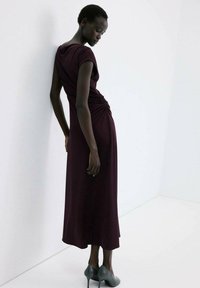 Vrouw in een donkerpaarse geplooide midi-jurk en grijze hoge hakken staat leunend tegen een witte muur met het hoofd naar beneden gekanteld.