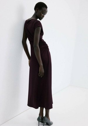 Mujer con un vestido midi fruncido de color púrpura oscuro y tacones altos grises, de pie, apoyándose ligeramente contra una pared blanca con la cabeza inclinada hacia abajo.