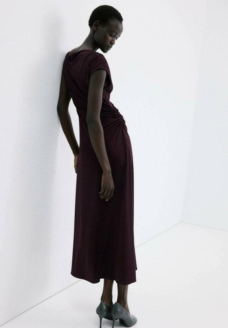 Vrouw in een donkerpaarse geplooide midi-jurk en grijze hoge hakken staat leunend tegen een witte muur met het hoofd naar beneden gekanteld.