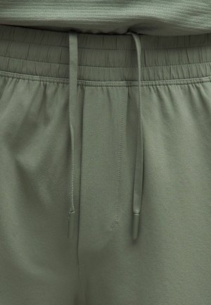 Elastyczny pas z dwoma zwisającymi troczkami na oliwkowozielonych casualowych spodniach, z widocznym szczegółowym przeszyciem i gładką teksturą tkaniny.