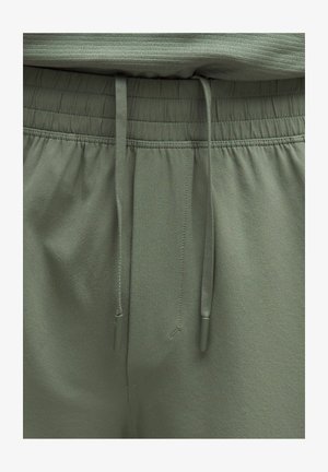 Elastyczny pas z dwoma zwisającymi troczkami na oliwkowozielonych casualowych spodniach, z widocznym szczegółowym przeszyciem i gładką teksturą tkaniny.