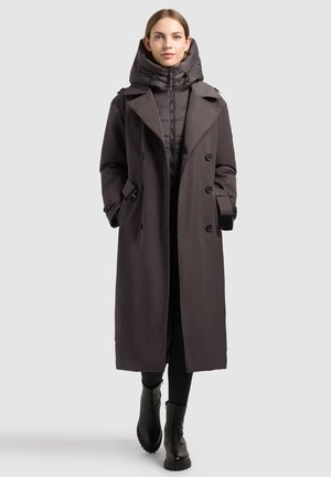 Femme portant un long manteau sombre par-dessus une veste matelassée avec capuche, leggings noirs et bottines noires, debout devant un fond uni.