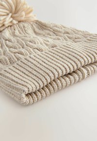 Bonnet en tricot beige clair avec un motif en cable et un revers en côtes. Présente un pompon duveteux sur le dessus. Fil doux et texturé utilisé.